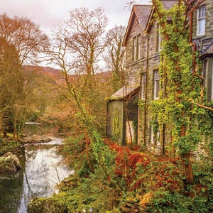 A stone building with large windows is surrounded by lush greenery and ivy next to a tranquil river with rocks and reflecting trees.