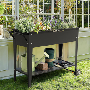 A black metal raised garden bed cart with a 38-inch width, featuring wheels and a lower shelf for storage. It holds various potted plants, gardening tools, a watering can, and bricks, positioned next to a greenhouse.