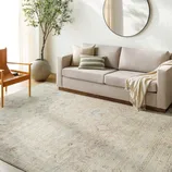 A light beige sofa with cushions and a throw blanket is positioned on a patterned area rug. A wooden chair and a large potted plant adorn the minimalist living space, which features a round wall mirror.