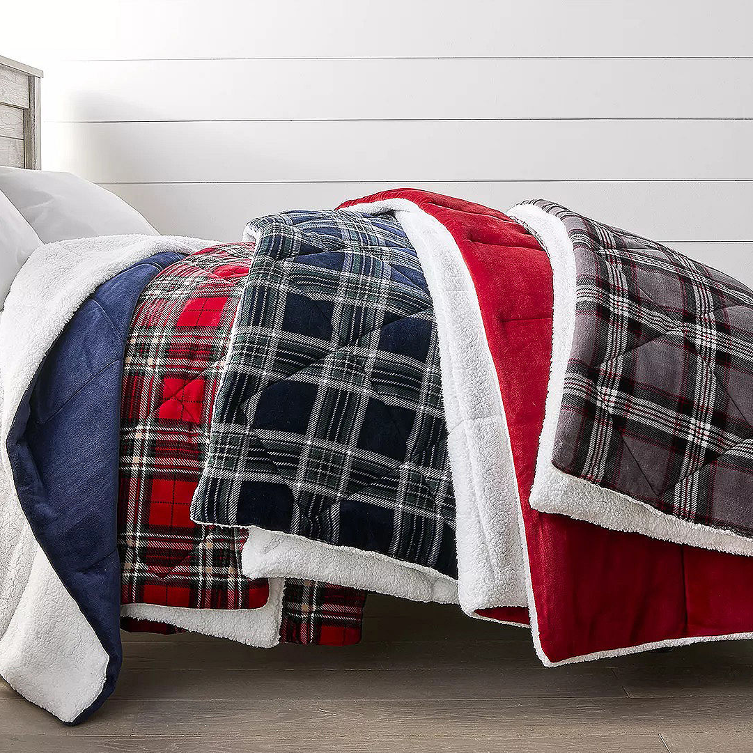 Quilted blankets with sherpa lining in plaid and solid patterns, featuring red, navy, and gray colors, neatly layered on a bed.