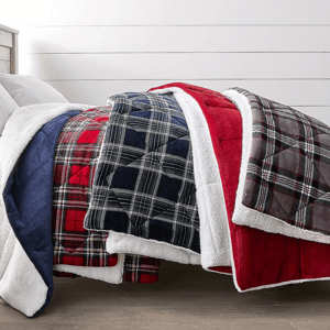 Quilted blankets with sherpa lining in plaid and solid patterns, featuring red, navy, and gray colors, neatly layered on a bed.