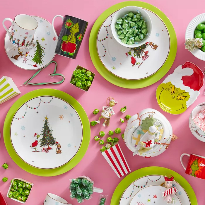 The Lenox Merry Grinchmas Collection features festive dinnerware including plates, mugs, and a tray with playful Grinch-themed designs in green and red. Accompanied by small candy dishes and mugs with Christmas motifs, all set against a pink background.