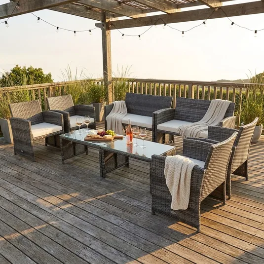 Outdoor patio setting with wicker furniture: a long glass-top table, six armchairs, a sofa, cushions, and draped blankets. A pergola with string lights and greenery in planters surrounds the setup.