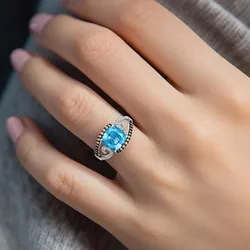 A hand with a silver ring featuring a large blue gemstone in the center, accented by smaller clear stones in a decorative setting.