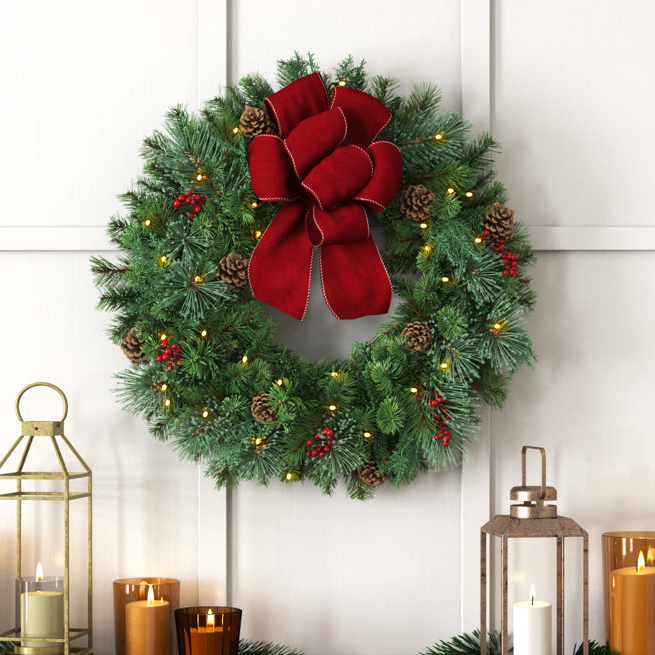 A Christmas wreath with a large red bow, lights, pine cones, and red berries hangs on a door, accompanied by candle lanterns on the side.