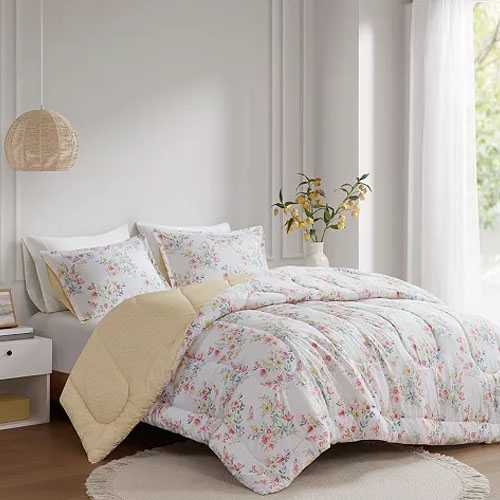 A bed with floral-patterned bedding and pillows, featuring a yellow underside, is displayed in a well-lit room with a round rug and a pendant lamp.