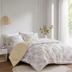A bed with floral-patterned bedding and pillows, featuring a yellow underside, is displayed in a well-lit room with a round rug and a pendant lamp.
