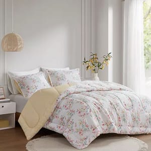 A bed with floral-patterned bedding and pillows, featuring a yellow underside, is displayed in a well-lit room with a round rug and a pendant lamp.