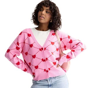 A woman is wearing a pink cardigan with red heart patterns over a white top, paired with light blue jeans and carrying a pink shoulder bag.