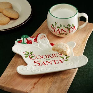 Cookies for Santa set featuring a Santa-themed plate and a mug for milk, both with holly decorations, on a wooden surface. A partially eaten cookie is on the plate, with a separate plate of cookies nearby.