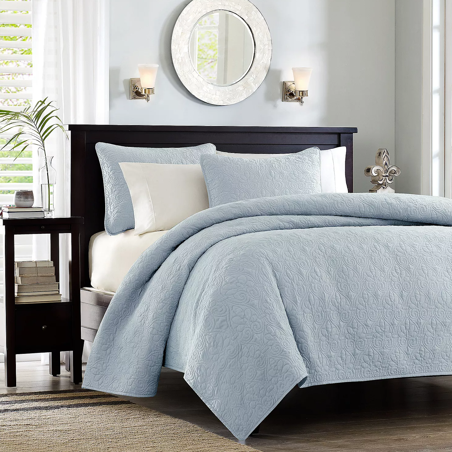A neatly made bed with a blue quilted bedspread and matching pillows against a dark headboard, flanked by a side table and wall sconces.