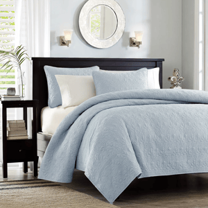 A neatly made bed with a blue quilted bedspread and matching pillows against a dark headboard, flanked by a side table and wall sconces.