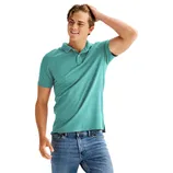 A person wearing a teal polo shirt and blue jeans stands casually with one hand touching their hair.