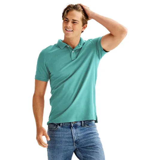 A person wearing a teal polo shirt and blue jeans stands casually with one hand touching their hair.
