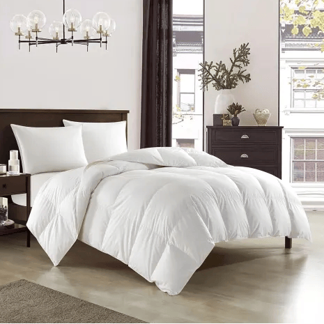 A bedroom features a plush white comforter on a bed with white pillows. The room includes a dark wood headboard, matching nightstand, a lamp, curtain, and a drawer chest with decorative items. A modern chandelier and large windows add brightness.