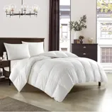 A bedroom features a plush white comforter on a bed with white pillows. The room includes a dark wood headboard, matching nightstand, a lamp, curtain, and a drawer chest with decorative items. A modern chandelier and large windows add brightness.