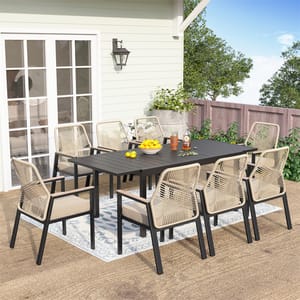 The 7pc Patio Dining Set includes a rectangular black table and six beige woven chairs with cushions, placed on an outdoor rug. The setup is situated on a patio next to a white-paneled wall with a sliding glass door and adjacent greenery.