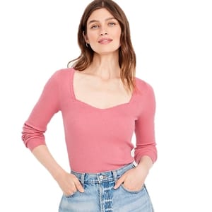 A woman is wearing a pink ribbed long-sleeve top with a sweetheart neckline and light blue high-waisted jeans.