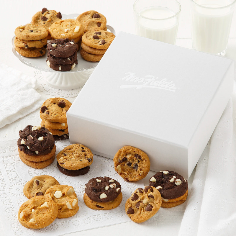 Assorted cookies, including chocolate chip and white chocolate varieties, are displayed alongside a white gift box labeled \“Mrs. Fields\“ and two glasses of milk.