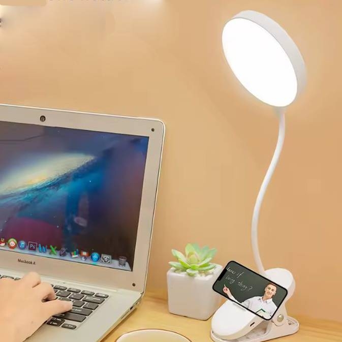 A laptop is placed on a desk beside a flexible desk lamp with a bright circular light. A smartphone is attached to the lamp's base, which also includes a small whiteboard with a person's image. A potted succulent is nearby.