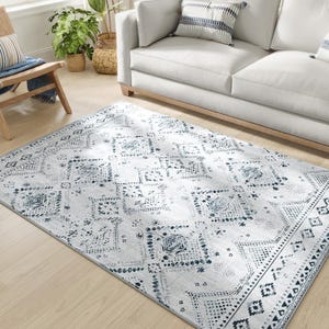 A 5' x 7' machine-washable area rug with a light gray base and intricate blue geometric and floral patterns, placed on a light wooden floor near a sofa and plant.