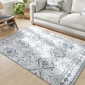 A 5' x 7' machine-washable area rug with a light gray base and intricate blue geometric and floral patterns, placed on a light wooden floor near a sofa and plant.