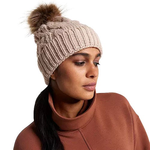 A beige cable-knit beanie with a ribbed texture and a fluffy pom-pom on top, worn by a woman with a ponytail sticking out from the back.