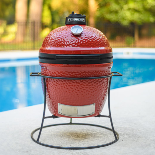 A red Kamado Joe Jr. charcoal grill with a dome thermometer, set on a black metal stand beside a pool, amidst a background of trees.