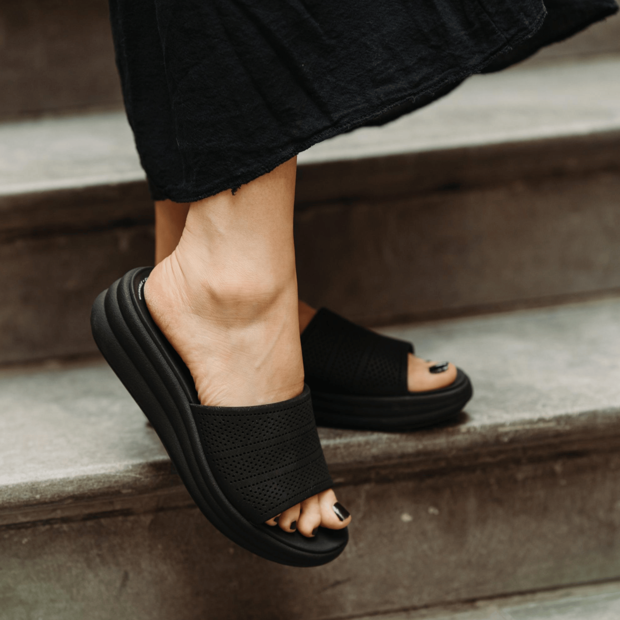 A person wears black slide sandals with a perforated strap and a thick, cushioned sole while standing on stairs.