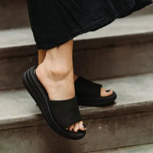 A person wears black slide sandals with a perforated strap and a thick, cushioned sole while standing on stairs.