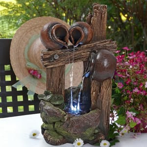 Decorative outdoor fountain featuring two rustic-style jugs with water flowing into a basin, illuminated by LED lights, set against a background of artificial wood and stone, alongside a straw hat and pink flowers.