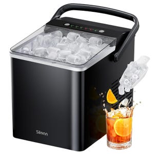 Black portable ice maker with a transparent lid, displaying clear ice cubes inside. It features a scoop on the side and is shown with a glass of iced beverage with orange slices.