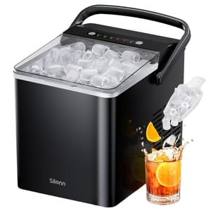 Black portable ice maker with a transparent lid, displaying clear ice cubes inside. It features a scoop on the side and is shown with a glass of iced beverage with orange slices.