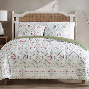 A bedding set featuring a white comforter and two pillow shams with a repeating floral pattern and subtle green accents. The bed has a woven cane headboard, and a wooden nightstand with a lamp is visible beside it.