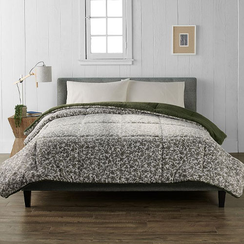 A bed with a gray upholstered headboard features a floral-patterned comforter and pillow set, alongside a green blanket. A wooden nightstand holds a lamp and a small plant, with a framed picture on the white paneled wall behind.