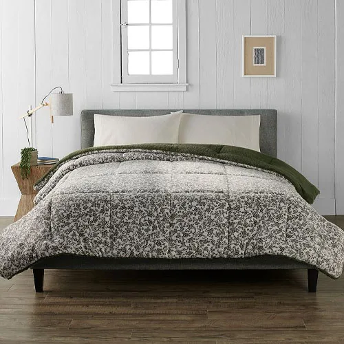 A bed with a gray upholstered headboard features a floral-patterned comforter and pillow set, alongside a green blanket. A wooden nightstand holds a lamp and a small plant, with a framed picture on the white paneled wall behind.