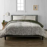 A bed with a gray upholstered headboard features a floral-patterned comforter and pillow set, alongside a green blanket. A wooden nightstand holds a lamp and a small plant, with a framed picture on the white paneled wall behind.