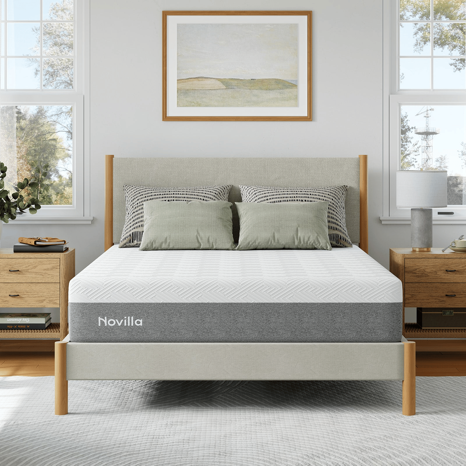 A modern bedroom featuring a Novilla mattress on a wooden bed frame, with two green and two patterned pillows. Matching wooden nightstands with a lamp and books complete the decor, enhanced by a framed landscape painting above the bed.