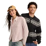 A woman in a striped button-up shirt, jeans, and a cap stands next to a man wearing a black and white patterned sweater.