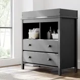 Gray changing table with two drawers and open shelves, featuring a basket with diapers and bottles.