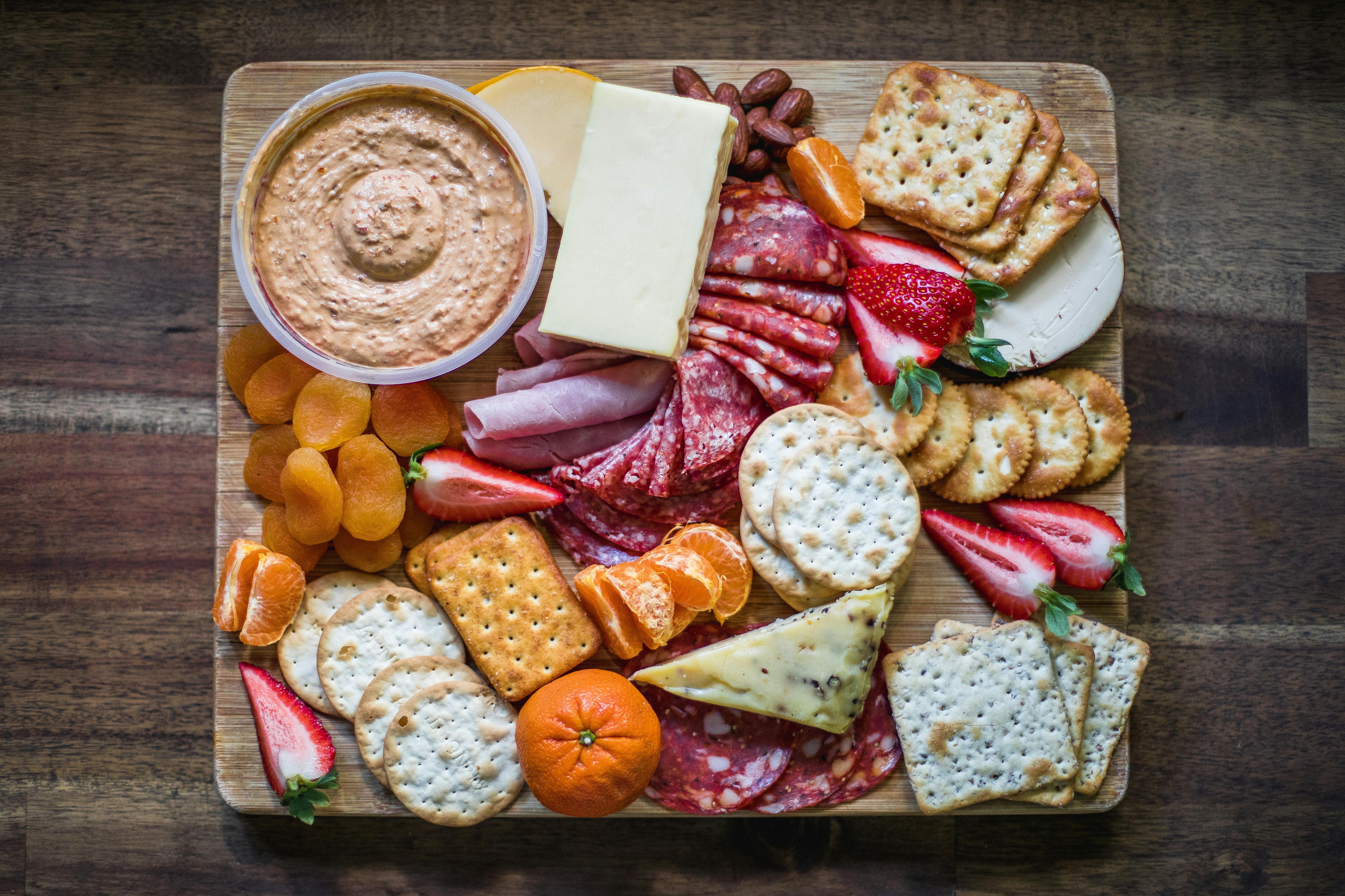 A charcuterie board featuring assorted meats, cheeses, crackers, dried apricots, strawberries, and clementines, with a bowl of dip.