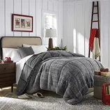 A cozy bedroom features a plush gray comforter, neutral pillows, a wooden nightstand with a lamp, and seasonal decor. A ladder with a red scarf leans against a white paneled wall, and a patterned rug and gift baskets complete the look.