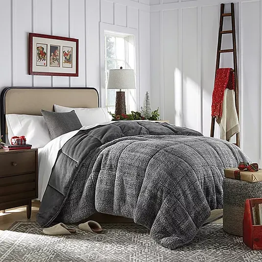 A cozy bedroom features a plush gray comforter, neutral pillows, a wooden nightstand with a lamp, and seasonal decor. A ladder with a red scarf leans against a white paneled wall, and a patterned rug and gift baskets complete the look.