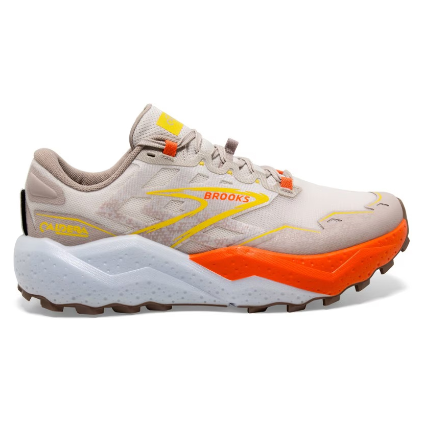 Brooks Caldera 7 Trail Shoes feature a beige upper with orange and yellow accents, a thick white and orange midsole for cushioning, and a durable brown outsole. Designed for trail running, they offer support and grip on various terrains.