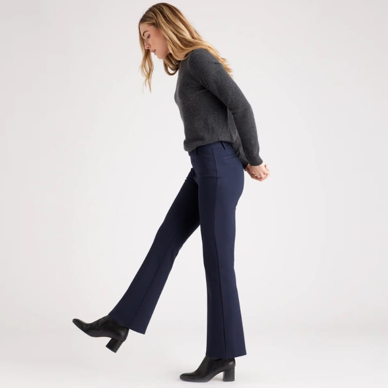 A woman is wearing a grey sweater, navy flared trousers, and black heeled boots.