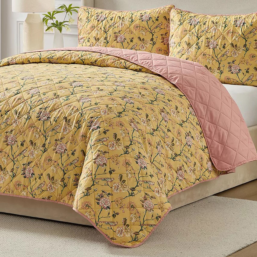 A quilt set with a floral pattern on a yellow background and a reversible pink backing, displayed with matching pillow shams on a bed.