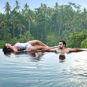 A couple relaxes on an infinity pool's edge, surrounded by lush palm trees, suggesting a luxurious and serene Bali suite stay.