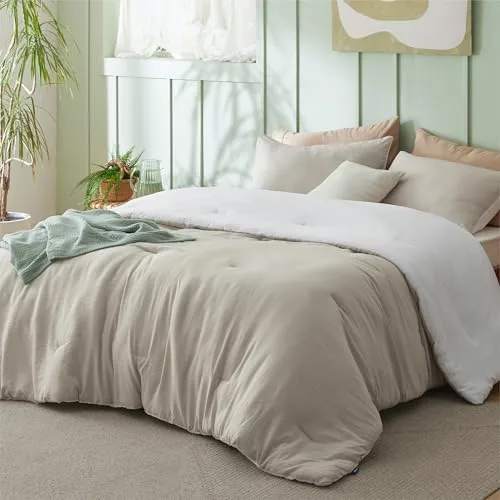 A cozy bed setup with a beige comforter, white blanket, and assorted pillows in a softly lit room featuring pastel green walls, a houseplant, and natural accents.