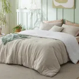 A cozy bed setup with a beige comforter, white blanket, and assorted pillows in a softly lit room featuring pastel green walls, a houseplant, and natural accents.