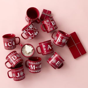 A collection of red holiday-themed mugs with individual letters on them, arranged next to two red gift boxes.